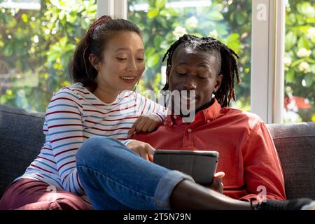 Allegri coppie diverse che usano il tablet seduto sul divano sorridendo nel soleggiato soggiorno Foto Stock