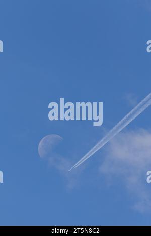 Una perfetta giornata estiva di sole con un aereo e sentieri di condensazione diretti verso una luna visibile in lontananza Foto Stock