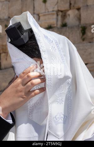 Un uomo ebreo che indossa i tefillin recita la preghiera Shema Yisrael durante il servizio Shacharit mentre nasconde il suo volto e gli occhi con il suo tallit o scialle di preghiera Foto Stock