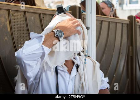 Un uomo ebreo che indossa i tefillin recita la preghiera Shema Yisrael durante il servizio Shacharit mentre nasconde il suo volto e gli occhi con il suo tallit o scialle di preghiera Foto Stock