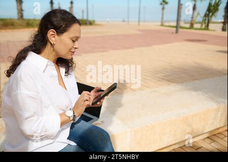 Giovane donna latinoamericana con smartphone che controlla i contenuti dei social media all'aperto. Donna d'affari in multitasking sicura che utilizza telefono cellulare, messaggistica, Foto Stock