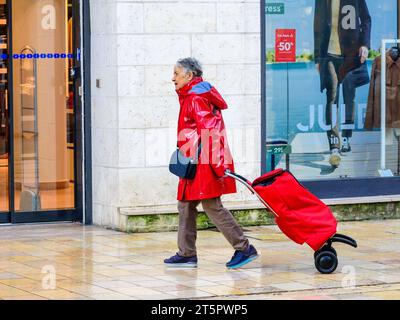 Donna anziana in plastica rossa impermeabile che tira il carrello rosso della spesa lungo la pavimentazione della città - Tours, Indre-et-Loire (37), Francia. Foto Stock