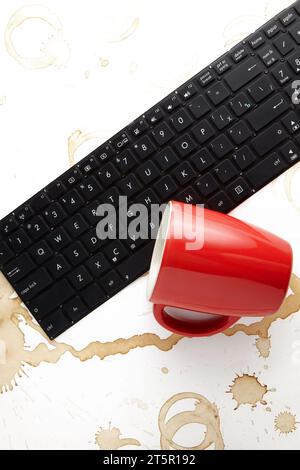 Una tastiera del computer portatile rovesciata e una tazza di caffè rossa rovesciata su uno sfondo bianco con macchie di caffè. Foto Stock