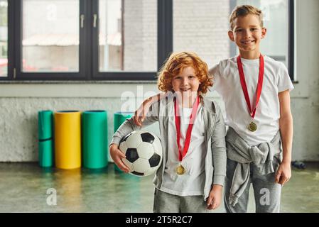 due ragazzi allegri in abbigliamento sportivo con medaglie d'oro che tengono la palla da calcio e sorridono alla macchina fotografica, sport Foto Stock