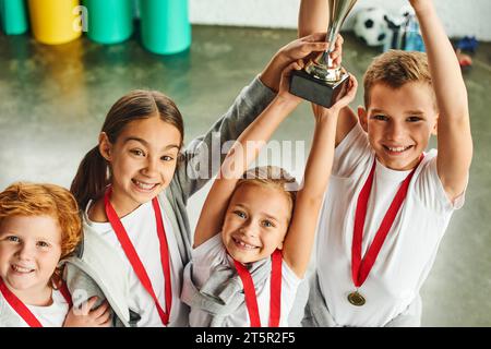 quattro simpatici bambini preadolescenti in abbigliamento sportivo trionfano con un trofeo enorme in mano Foto Stock