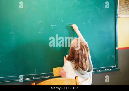 La piccola studentessa scrive sulla lavagna scolastica con gesso in classe. Bambino che risolve semplici problemi di matematica. Di nuovo a scuola e all'istruzione Foto Stock