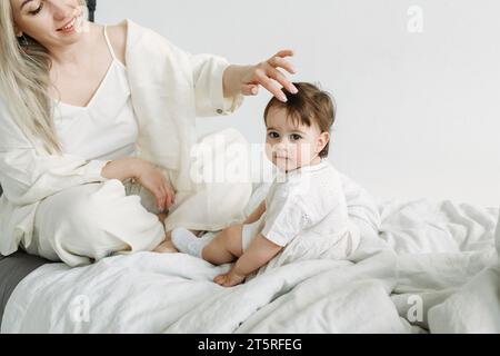 Ritratto di una bambina seduta accanto a sua madre sul letto la mattina dopo il sonno. Mamma raddrizza una ciocca di capelli per sua figlia. Co. Famiglia Foto Stock