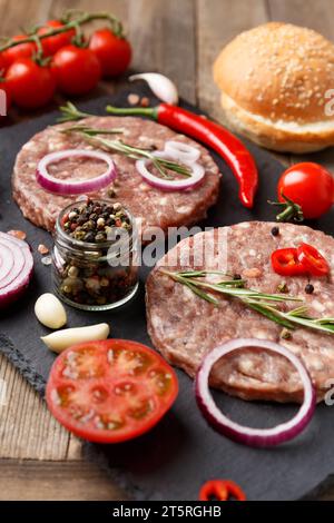 Hamburger di manzo macinato crudo, rosmarino, verdure e spezie su un piatto di pietra nera su uno sfondo di legno. Ingredienti per un hamburger. Foto Stock