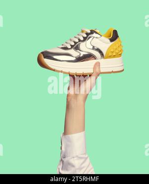 La mano di una donna, che indossa una camicia bianca, che regge un'elegante sneaker multicolor con una suola pesante, inserti in argento lucido e un bancone giallo con punte, Foto Stock