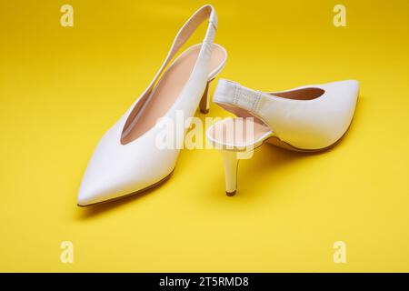 Eleganti scarpe da donna in pelle bianca con tacco alto e apertura sul retro, isolate su uno sfondo giallo brillante. Vista ravvicinata. Femminile alla moda Foto Stock