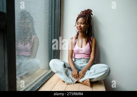 Attraente donna africana sorridente che indossa una canotta a coste e jeans seduto a gambe incrociate sul davanzale e guarda attraverso la finestra. Vita urbana c Foto Stock