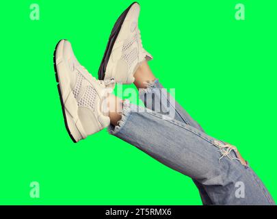 Foto ritagliata delle gambe di donna in eleganti sneaker bianche e jeans blu strappati, isolati su uno sfondo verde vivace. Design moderno, layout creativo. Foto Stock