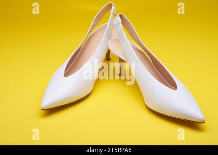 Eleganti scarpe bianche da donna con tacco alto e apertura sul retro con spalline posteriori, isolate su uno sfondo giallo brillante. Vista ravvicinata. Elegante e alla moda Foto Stock