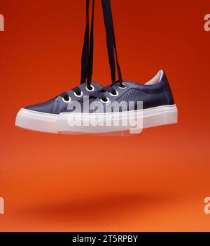 Sneaker eleganti e casual blu navy appese di fronte a uno sfondo arancione brillante. Pelle traforata, suola bianca spessa, lacci larghi. Scarpe casual, Foto Stock