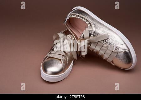 Sneaker eleganti in metallo dorato con dettagli in pelle stropicciata e suola in gomma bianca su sfondo marrone sfumato. Vista ravvicinata, una scarpa si inclina Foto Stock