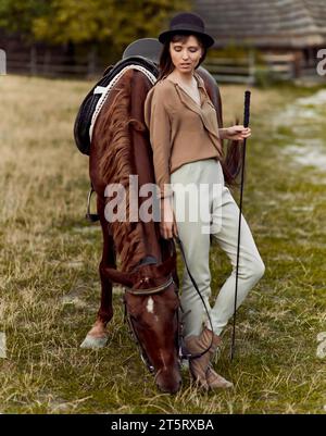 Una giovane donna con un cappello nero sta pascolando un cavallo marrone. Una donna e il suo cavallo in campagna, su un campo erboso. Foto Stock