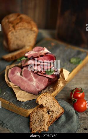 Un grande pezzo di prosciutto appetitoso si trova su una pergamena su una tavola di legno, con un pezzo di pane di segale e due pomodori davanti. Fotografia del cibo. Aestheti Foto Stock