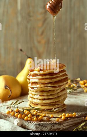 Una pila di pancake al burro disposti ad arte su una tavola di legno con un ciuffo di spina dorsale, il miele scorre da un cucchiaio di legno. Due pere forniscono S Foto Stock