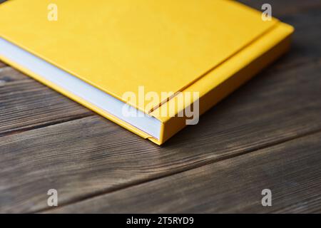 Fotobook giallo con copertina rigida isolato su uno sfondo di legno con spazio per le copie. Vista in prospettiva. Modello a libro chiuso vuoto per inserire testo personalizzato o immagine Foto Stock