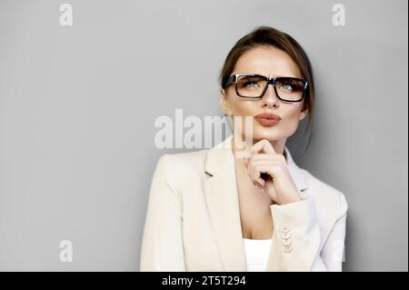 L'attraente donna pensiva con giacca beige e occhiali che guarda in alto tocca il mento con la mano su uno sfondo grigio con spazio per le copie. Tariffa donna d'affari Foto Stock