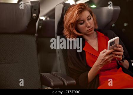 Giovane bella donna che viaggia in treno seduto vicino alla finestra utilizzando uno smartphone mobile. Stanco per un lungo viaggio. Lettura su cellulare social m Foto Stock