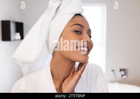Ritratto di felice donna birazziale che indossa accappatoio bianco e tocca la pelle in bagno Foto Stock