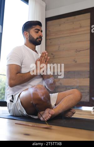 Rilassato uomo birazziale seduto, praticando yoga chiudendo gli occhi e meditando in una stanza soleggiata a casa Foto Stock