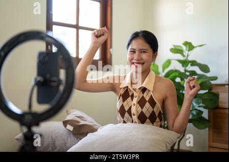 Una bella e allegra influencer o blogger asiatica sta parlando con la telecamera, registrandosi o trasmettendo in diretta sul suo social med Foto Stock