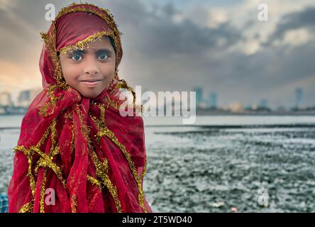 Una giovane, graziosa e sorridente ragazza mendicante musulmana fuori Haji Ali Dargah e la Moschea a Worli, Mumbai, India, strettamente coperta da un vestito rosso contro il vento Foto Stock