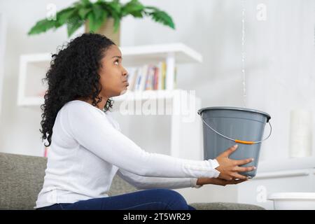 Preoccupato della donna tenendo la benna mentre le goccioline di acqua perdite dal soffitto Foto Stock