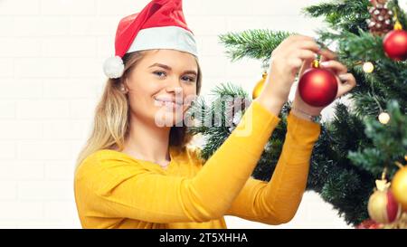 Giovane donna felice in un accogliente maglione che decora l'albero di natale con bauble a casa Foto Stock