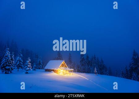 Un paesaggio invernale caratterizzato da una cabina isolata in legno e abeti innevati su un prato di montagna nel profondo della foresta. Cartolina di Natale. Foresta di montagne innevate Foto Stock