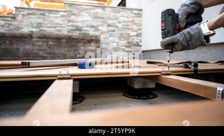 Progetto fai-da-te di posa di una terrazza esterna in legno - vista ad angolo basso delle mani maschili in guanti protettivi utilizzando un cacciavite elettrico per fissare le tavole in legno Foto Stock