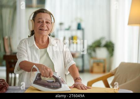 Ritratto di una donna anziana allegra che ascolta audiolibro in cuffia mentre stirano i vestiti Foto Stock