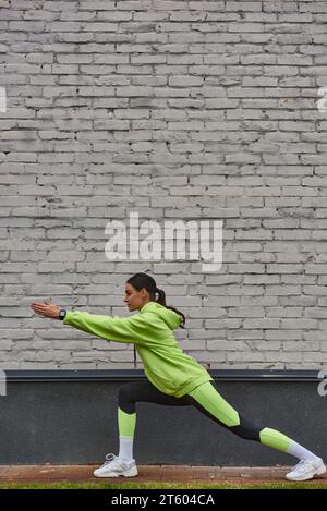 vista laterale di una donna sportiva con cappuccio e leggings di colore calce che fa affondi prima di correre all'aperto Foto Stock