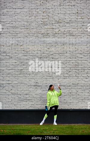 felicità sportiva con felpa e leggings che parla di foto sullo smartphone e tiene in mano una bottiglia d'acqua Foto Stock