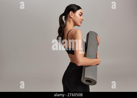 vista laterale, donna sportiva con top ritagliato e leggings con tappetino fitness e in piedi su sfondo grigio Foto Stock