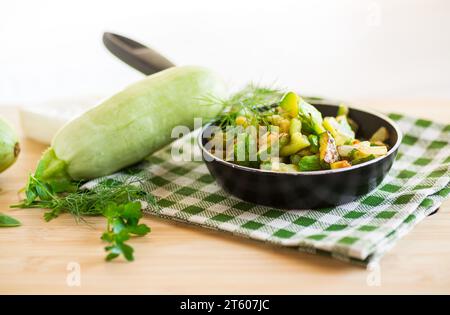 Zucchine a fette fritte con cipolle ed erbe in una padella su un tavolo di legno. Foto Stock