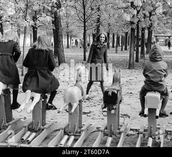 Anni '1960, la modella donna guarda i bambini che giocano su una giostra a cavallo, il giardino Jardin des Tuileries, Parigi, Francia, Europa, Foto Stock