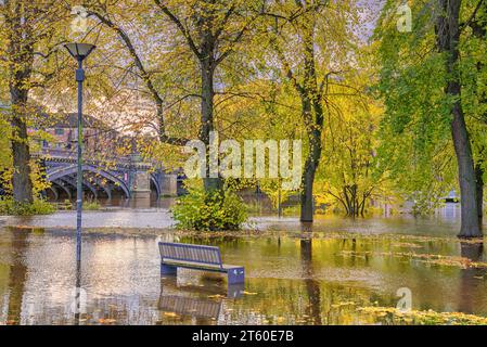 Un fiume ha scoppiato le sue rive e allagato attraverso un parco. Alberi autunnali e un lampione sono bloccati nell'acqua e una panca è parzialmente sommersa. Foto Stock