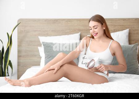 Giovane donna che depone le gambe con un moderno fotoepilatore in camera da letto a casa Foto Stock