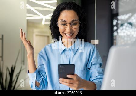Felice donna ispanica di successo che sorride leggendo la notifica, celebrando il successo della vittoria, donna d'affari ha ricevuto un messaggio online di notizie, tenendo in mano il telefono, utilizzando l'app sullo smartphone. Foto Stock