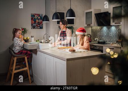 Graziose bambine con i cappelli rossi di Babbo Natale con la madre che prepara biscotti fatti in casa al pan di zenzero usando i taglierini per biscotti insieme nella cucina casalinga Foto Stock