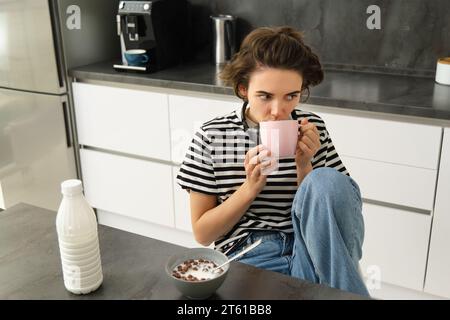 Ritratto di una donna moderna ed elegante, beve tè, sorseggia dalla sua tazza, siede sulla sedia in cucina, a fare colazione, cereali con latte Foto Stock