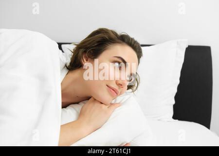 Ritratto ravvicinato di una bella bruna, che si sveglia nel suo letto, guarda la sveglia e guarda da parte con un volto sorridente e sognante Foto Stock