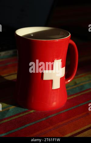 Tazza rossa con croce bianca svizzera isolata su una tovaglia colorata. Accogliente atmosfera invernale svizzera. Tazza da caffè con bandiera svizzera (rossa con croce bianca) Foto Stock