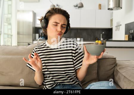 Giovane donna felice ed elegante, ascolta la musica in cuffia, mangia una ciotola di cereali, balla e si siede sul divano in salotto, facendo colazione Foto Stock