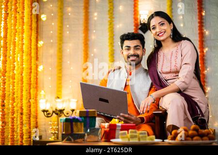 Felice e sorridente giovane coppia indiana che usa un computer portatile guardando la fotocamera mentre si siede sul divano a casa - concetto di offerte del festival, relazioni e. Foto Stock