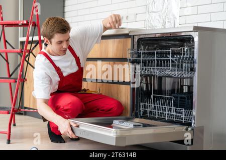 Il tecnico o il lavoratore in uniforme installa la lavastoviglie nei mobili della cucina. Il riparatore indossa tuta da lavoro riparando la manutenzione della lavastoviglie. Maestro Foto Stock