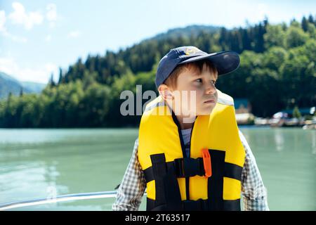 Un ragazzo di liitle che indossa un giubbotto di sicurezza si sente malato in barca a remi sullo splendido lago turchese. montagne sullo sfondo Foto Stock